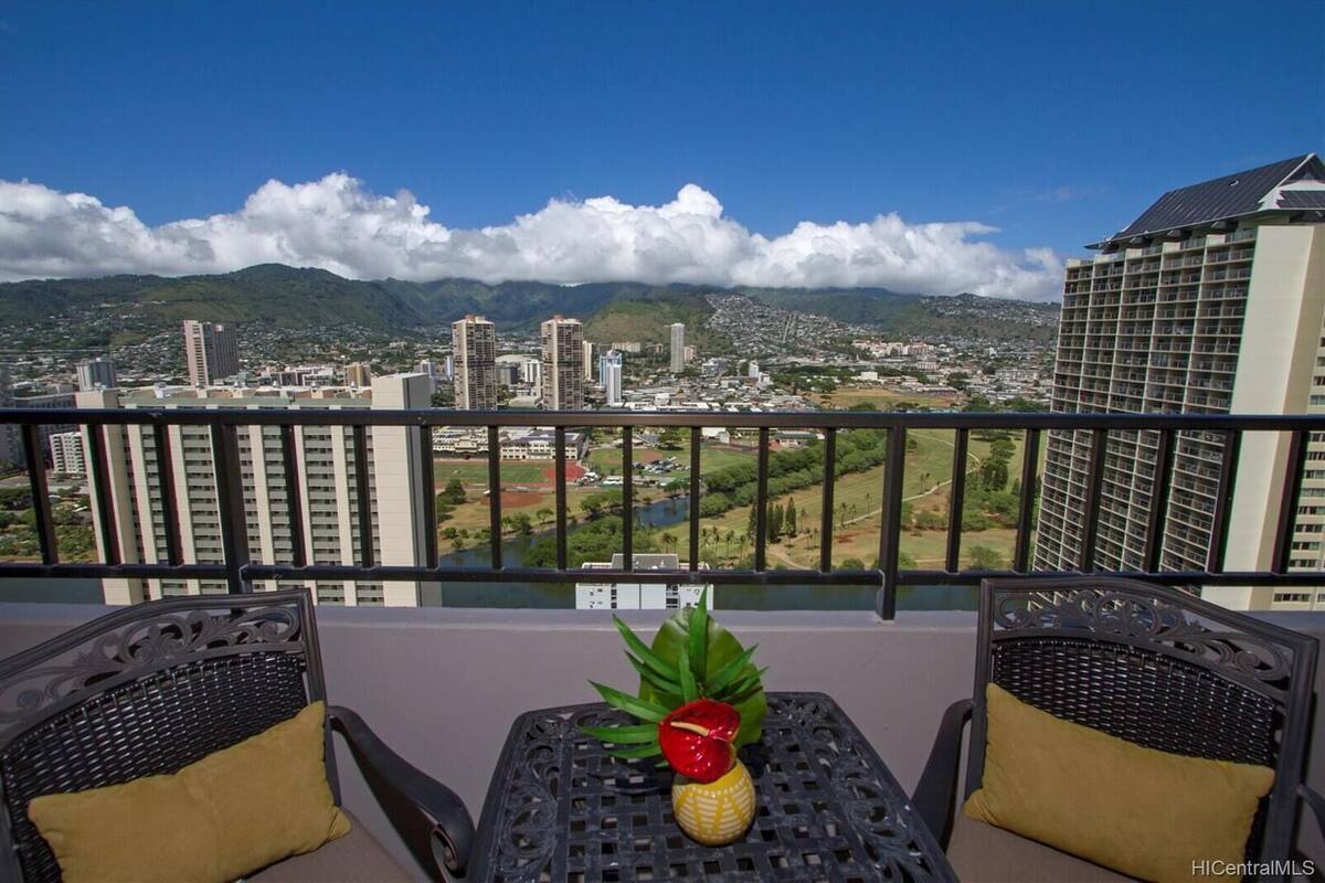 Un balcon avec deux fauteuils en osier, une petite table et une vue sur un paysage urbain, avec des montagnes en arrière-plan.