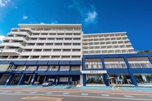 A multi-story hotel with a blue and white facade, multiple balconies, and a parking area in front.