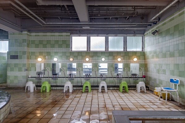 Un baño público con paredes de azulejos verde y blanco, varios lavamanos y taburetes de plástico verde y blanco.