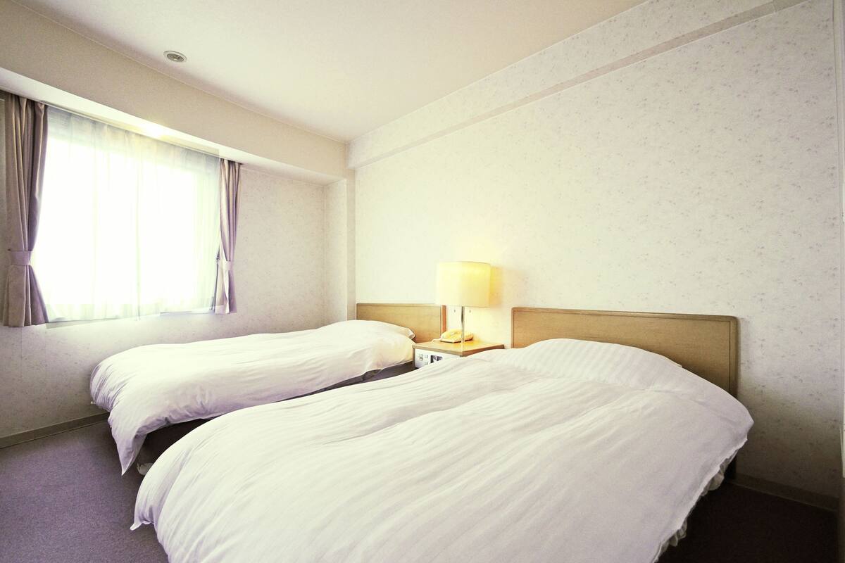 Hotellihuone, jossa on kaksi erillistä sänkyä, yöpöytä lampun kanssa ja ikkuna, jossa on verhot.