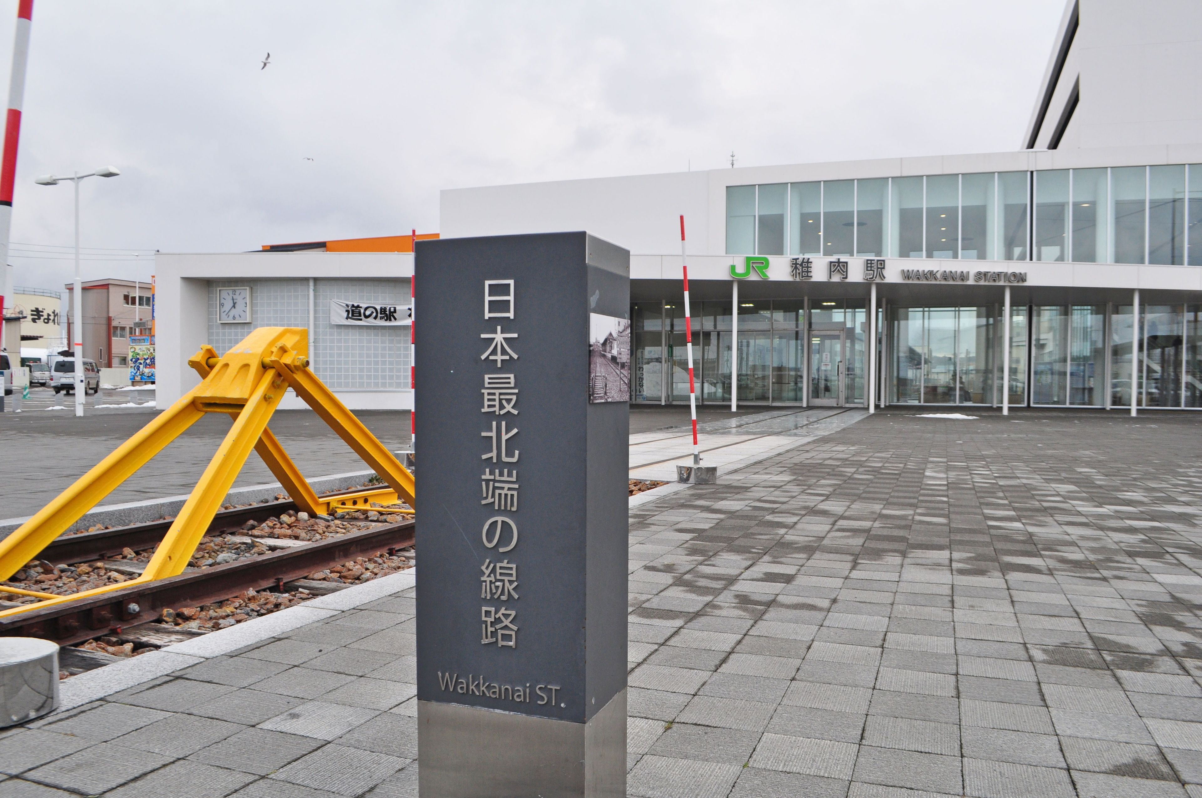 A train station entrance with a sign in Japanese, a modern building in the background, and a yellow safety barrier.