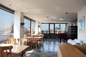 A spacious dining area with wooden tables and chairs, large windows offering a city view, and a balcony with a view of the sea. - Hotel Hachimanzaka (Hakodate)