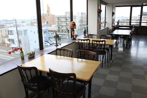 A restaurant with multiple tables and chairs, large windows with a city view, and various decorative items on the windowsill. - Hotel Hachimanzaka (Hakodate)