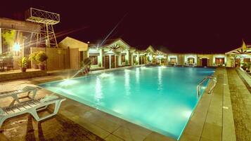 A swimming pool with a sunken seating area, surrounded by buildings with outdoor lighting.