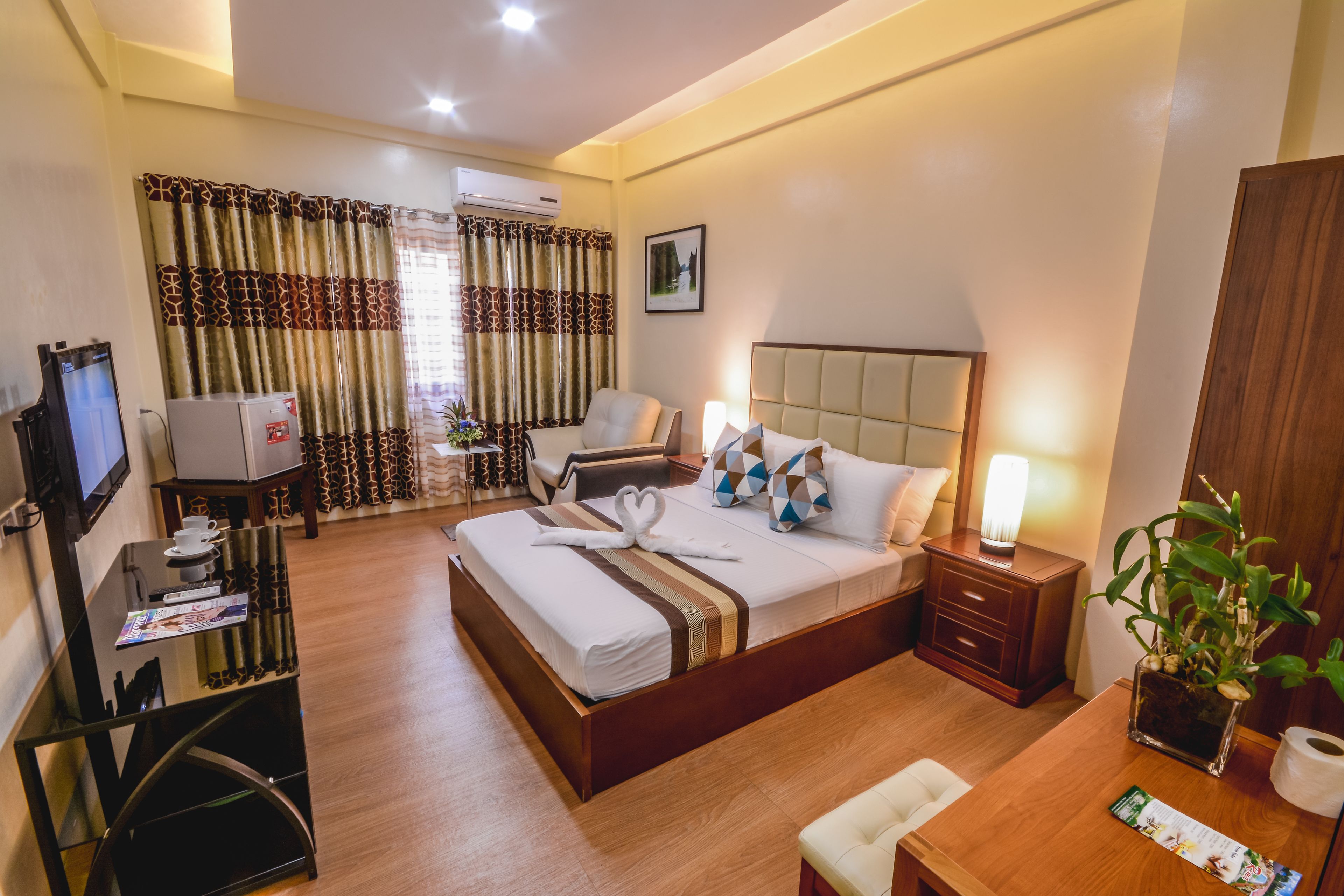 A hotel room with a bed, a desk, a TV, a chair, a nightstand, a lamp, and a plant.