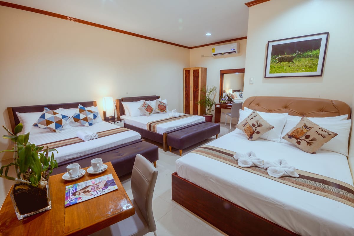 A hotel room with two beds, a wooden table with a magazine, a plant, and a framed picture on the wall.