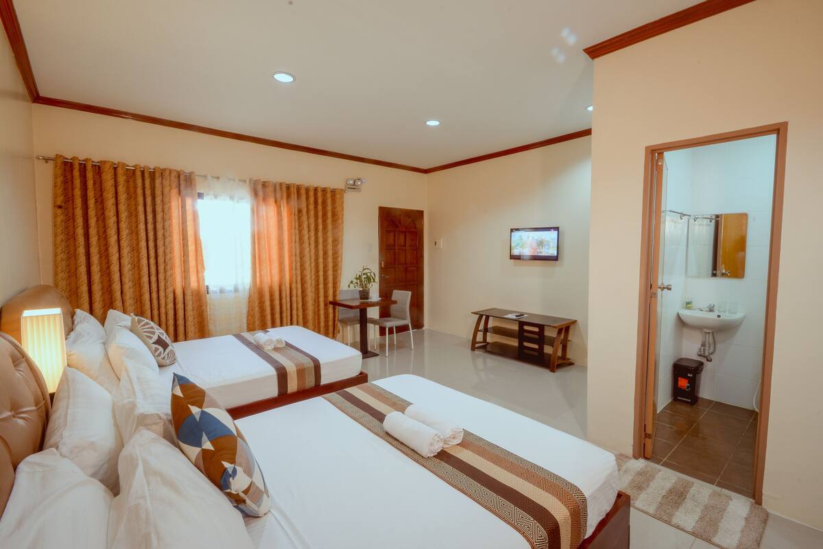 A hotel room with two beds, a desk, a chair, a TV, and a bathroom with a sink and mirror.