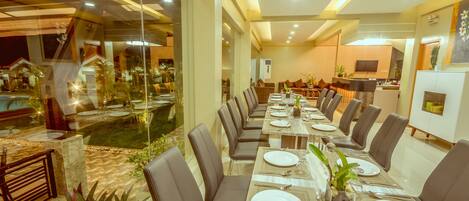 A long dining table with multiple chairs, a kitchen area, and a view of a pool and outdoor area.