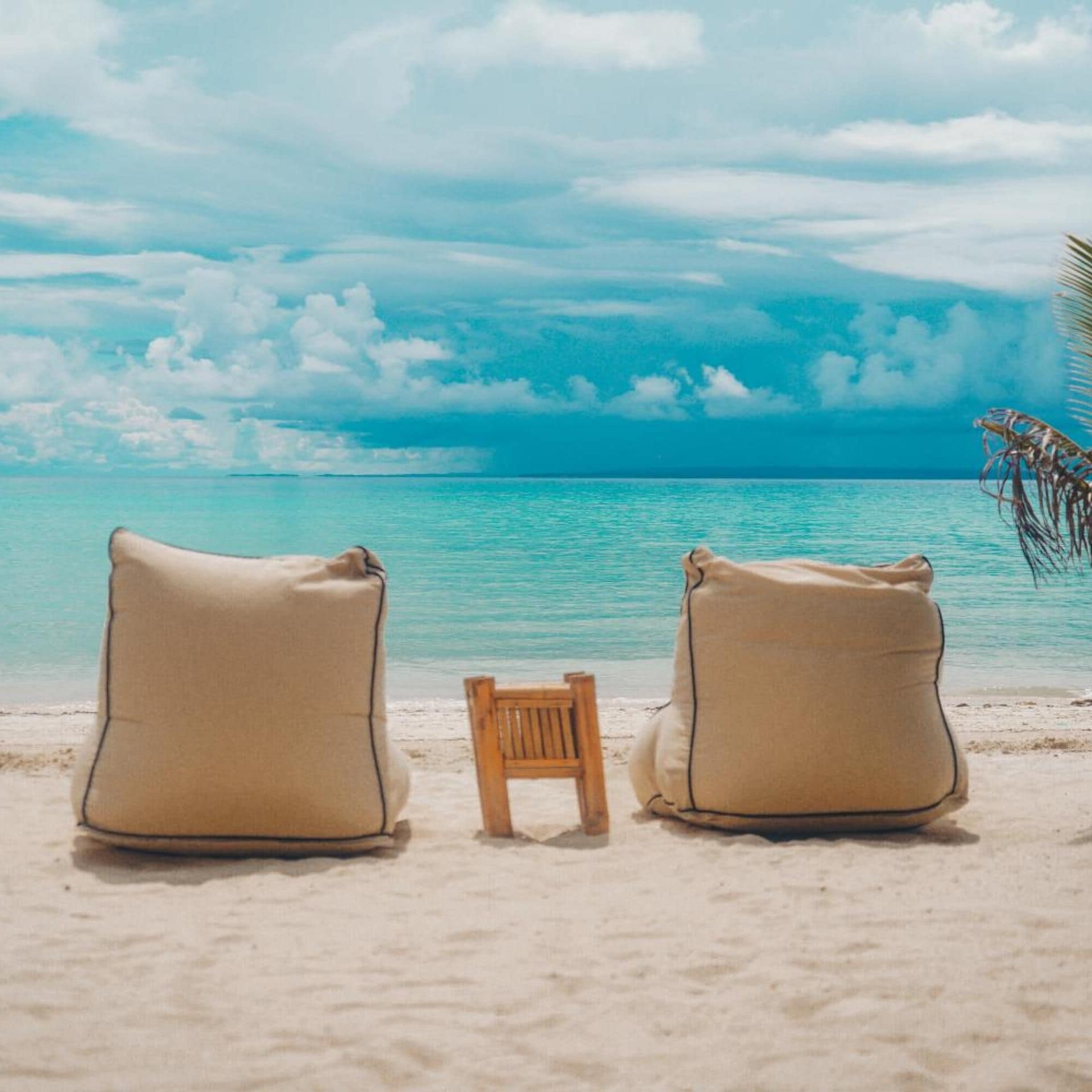 Two beige pillows on a sandy beach with a wooden chair in between, facing a calm turquoise sea under a partly cloudy sky.