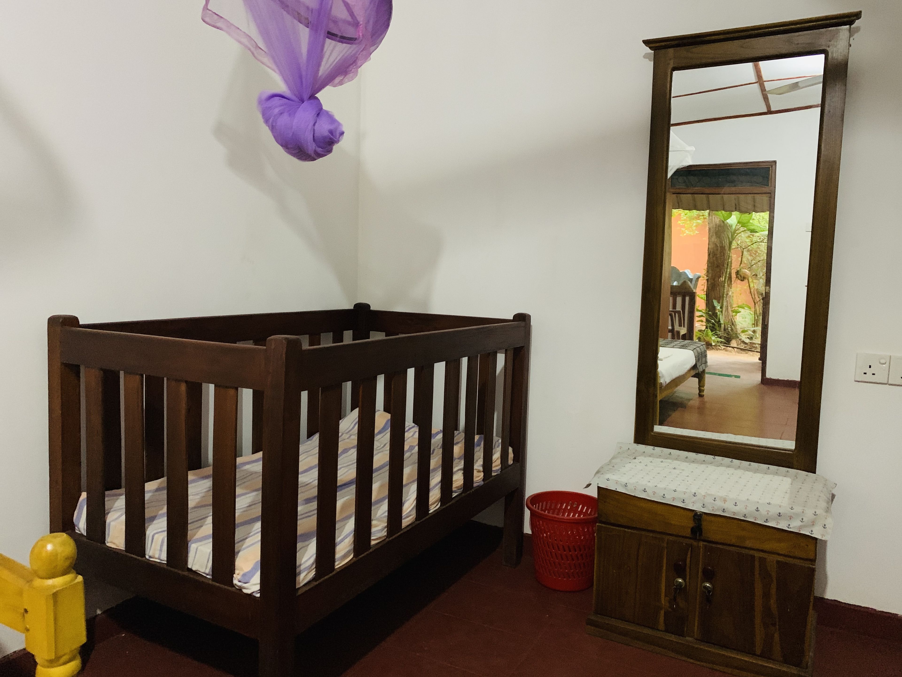 Una cuna de madera con colchón a rayas, un mueble de madera pequeño y un espejo en la pared.
