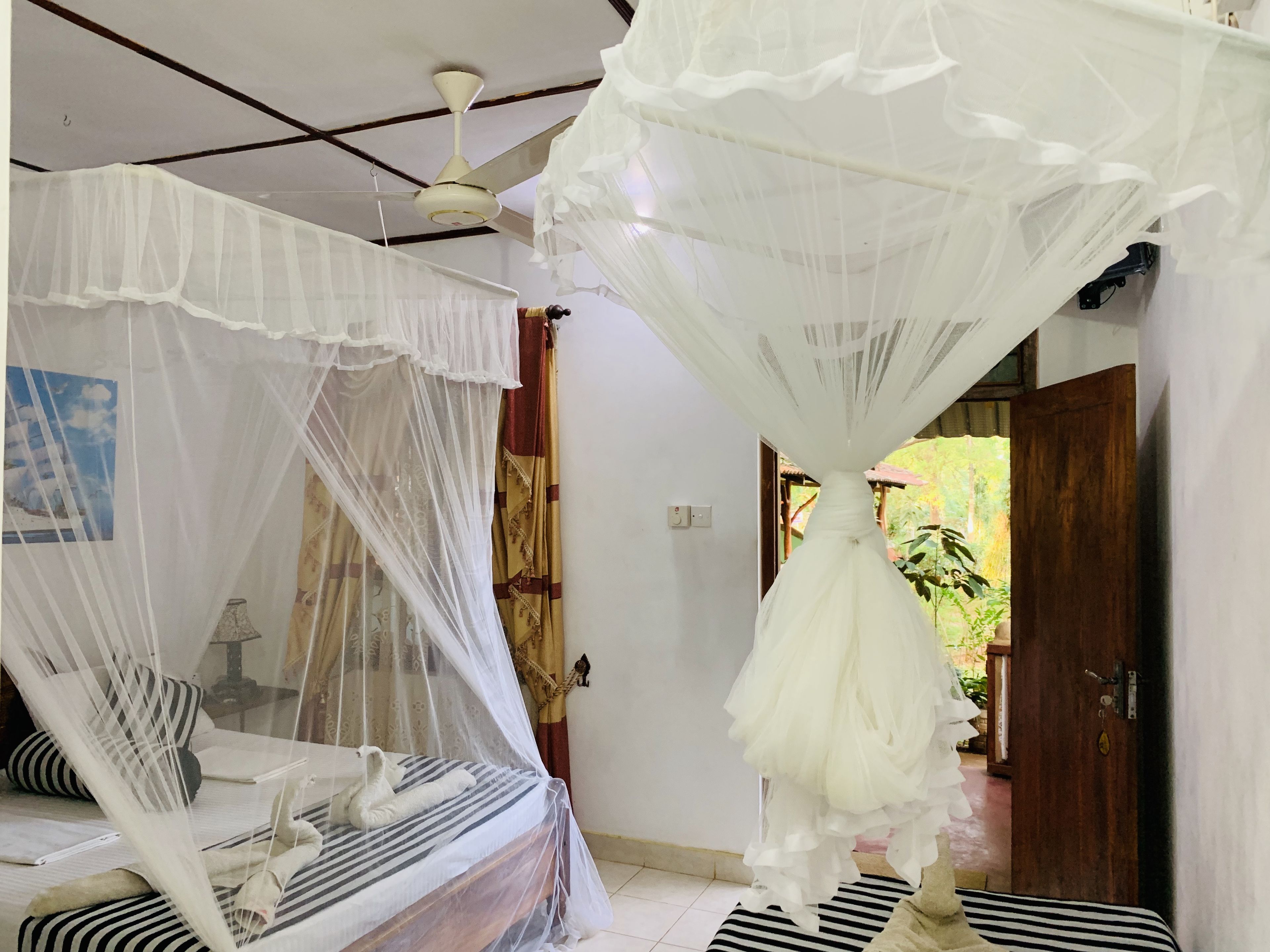A room with a bed under a white canopy, a ceiling fan, and a view of a garden through an open door.