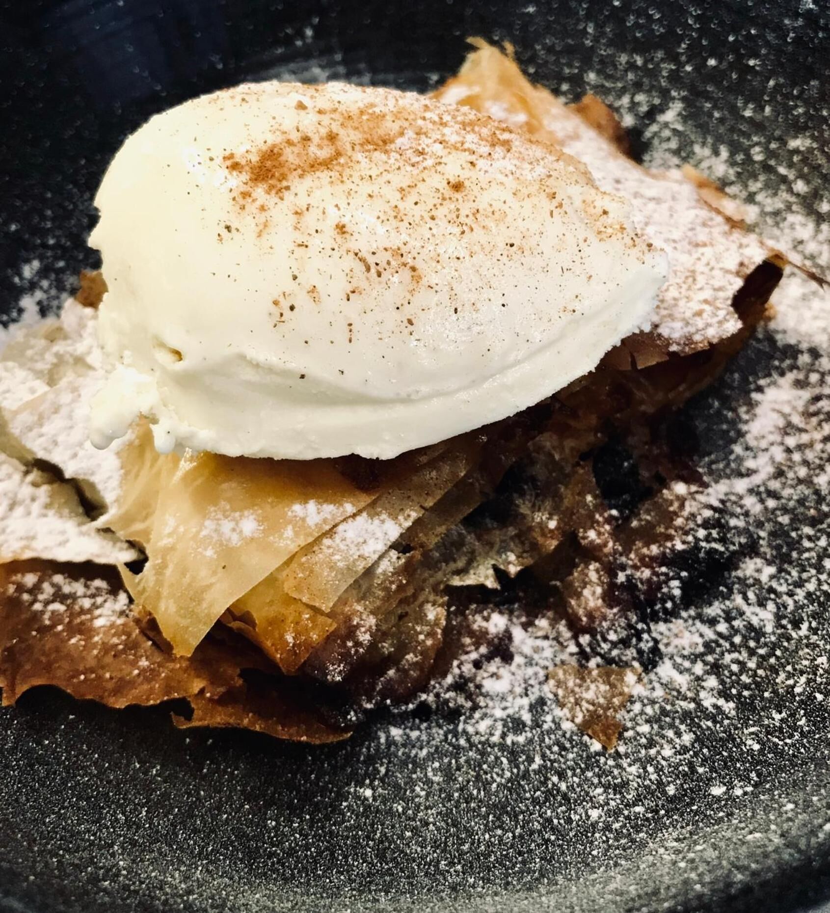 Dessert med en is kugle ovenpå, drysset med krydderi, serveret på en tallerken med mørk baggrund.