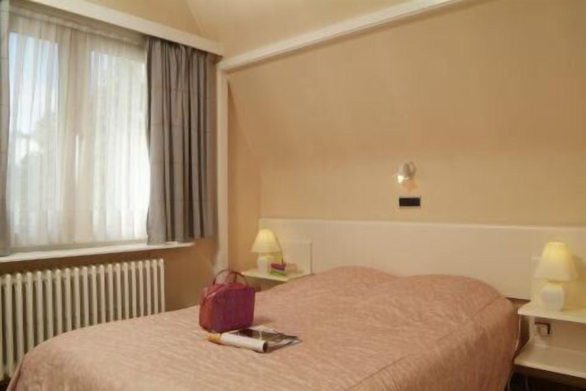 Une chambre d’hôtel avec un lit, une fenêtre avec des rideaux, un radiateur, une table de chevet avec une lampe et une petite table sur laquelle se trouvent un sac à main et un livre.