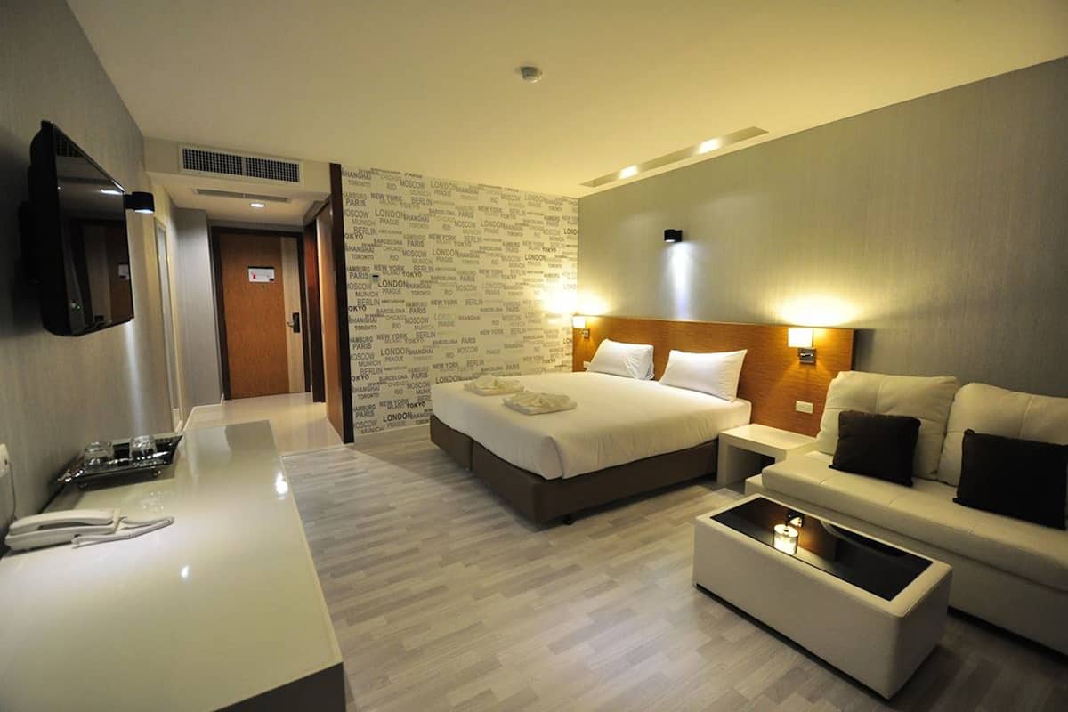 Quarto de hotel com cama, sofá, mesa de centro, escrivaninha com telefone e televisão na parede.