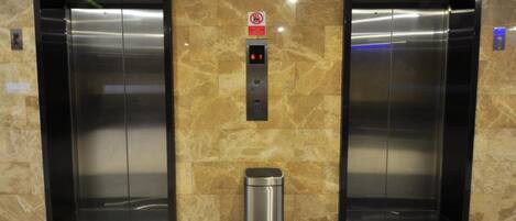 Two elevators with a digital display and a trash bin in a lobby with marble walls.