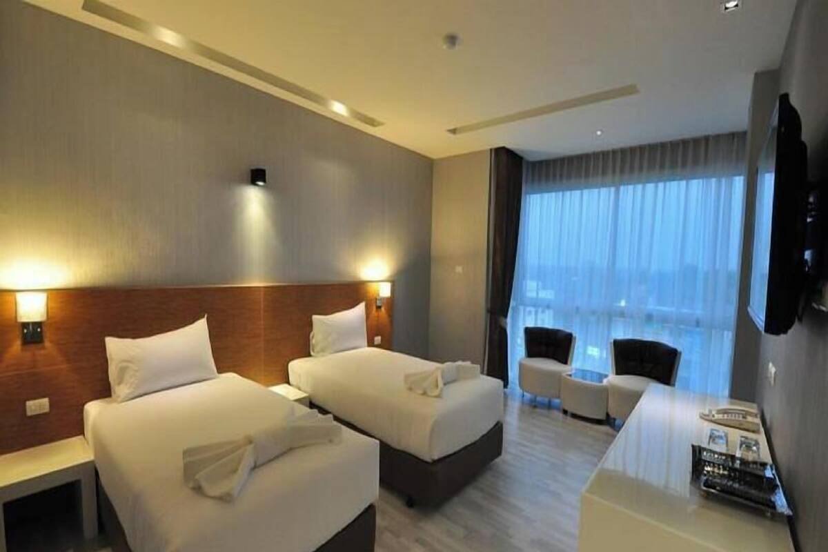 Quarto de hotel com duas camas, uma escrivaninha, uma cadeira e uma janela grande com cortinas.