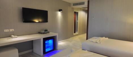 A modern hotel room with a flat-screen TV mounted on the wall, a desk with a phone and a small table, a bed with white bedding, and a small seating area with a bench.