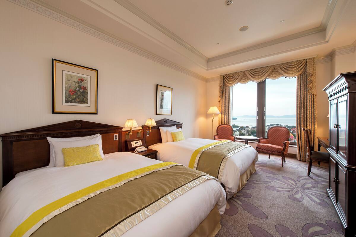 Habitación de hotel con dos camas, un escritorio, una silla, una lámpara y vistas a la ciudad.