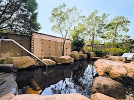 Un jardín tranquilo con un estanque de peces koi, grandes rocas, una valla de madera y una escalera metálica que lleva a una zona cubierta.