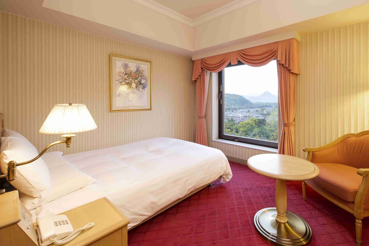 Habitación de hotel con cama, mesita de noche con lámpara, una silla, una pequeña mesa redonda, ventana con cortinas y un cuadro en la pared.