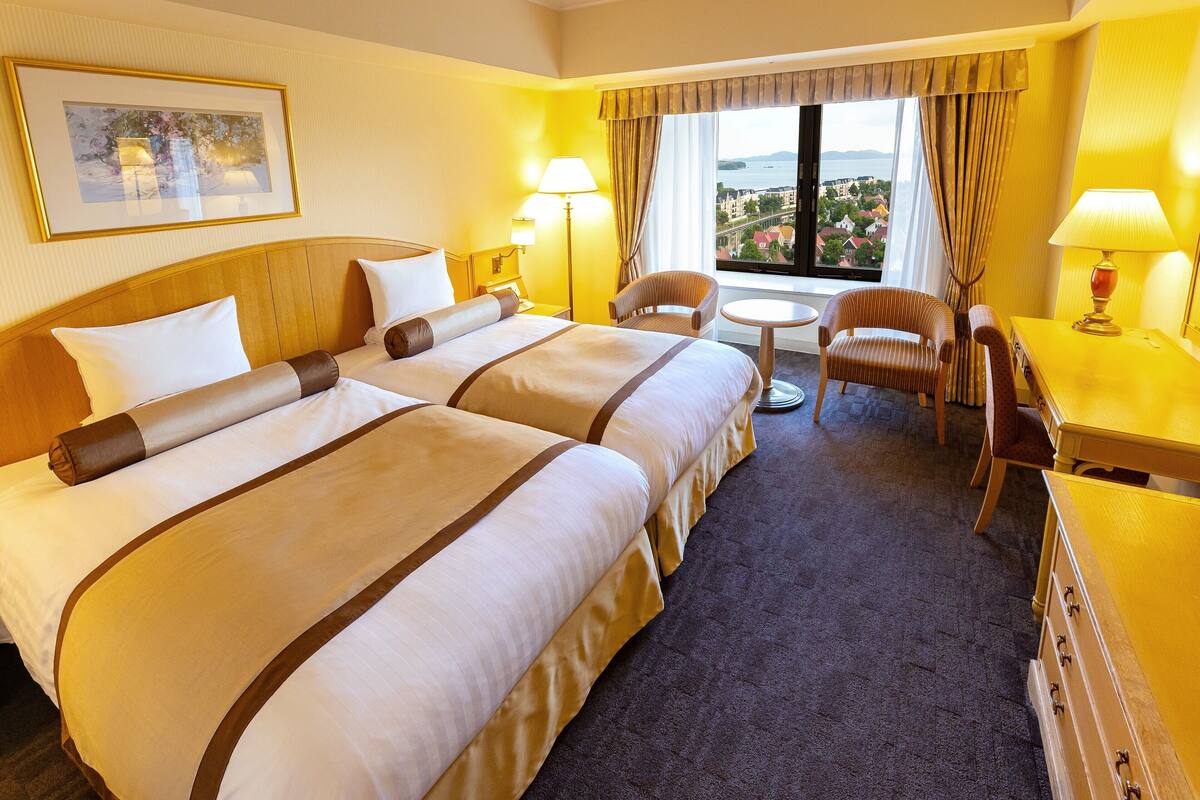 Habitación de hotel con dos camas, un escritorio, una silla, una lámpara y vista a la ciudad.