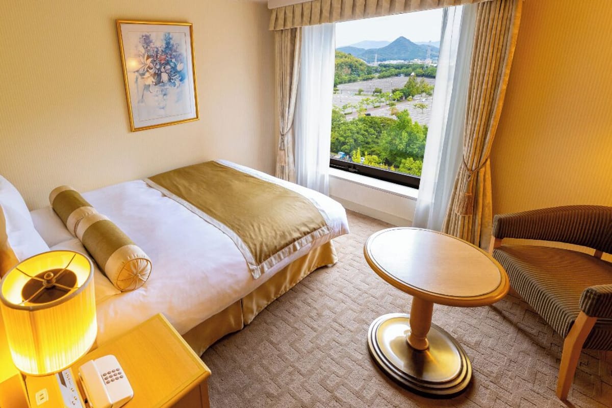 Habitación de hotel con cama, mesita de noche con lámpara, silla y vistas a zonas verdes y montañas a través del ventanal.