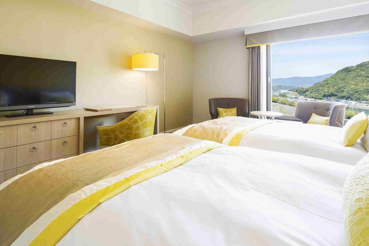 A hotel room with two beds, a TV, a desk, and a view of a mountainous landscape.