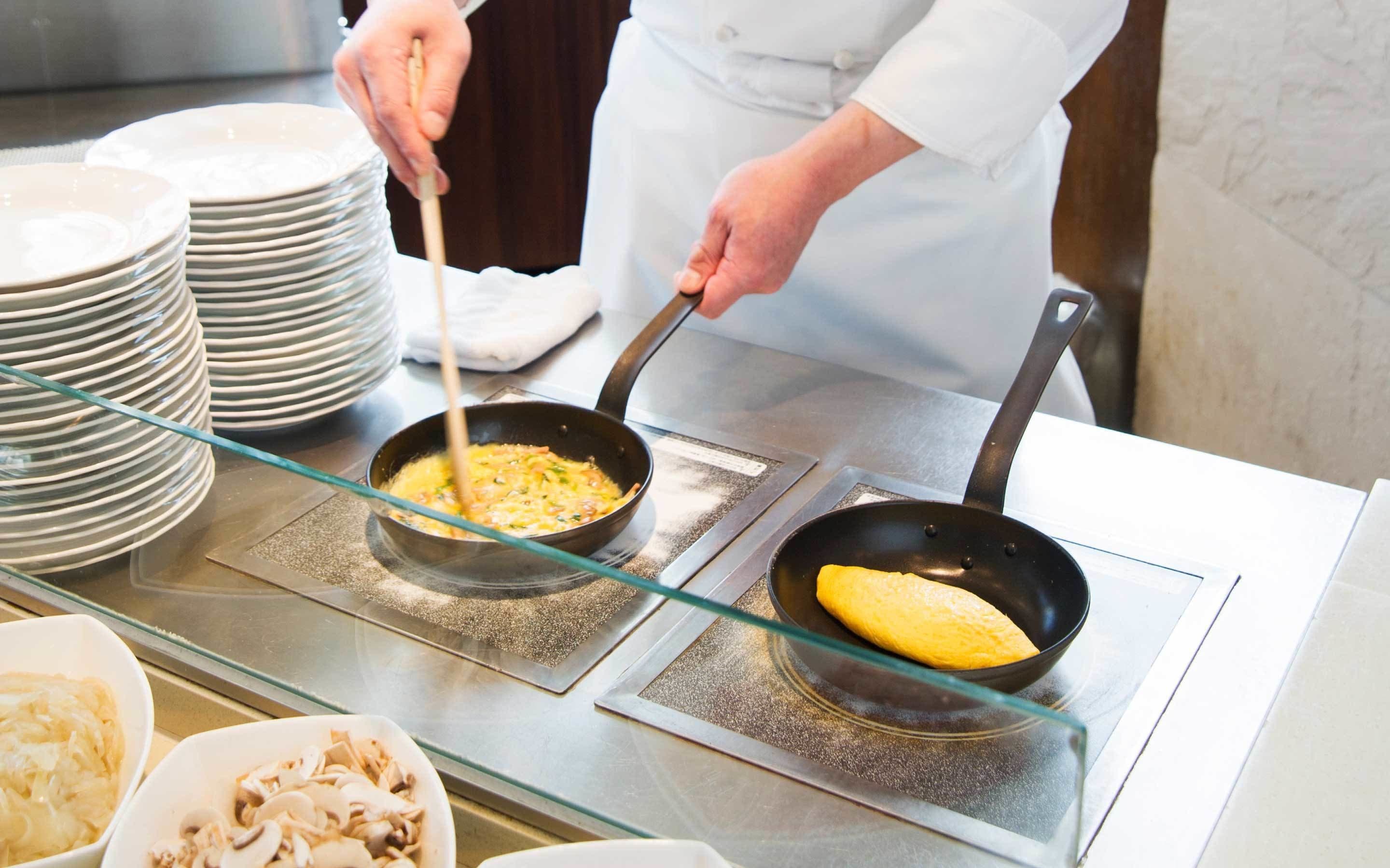 A kitchen with two frying pans on a stove, a stack of plates, and bowls of food.