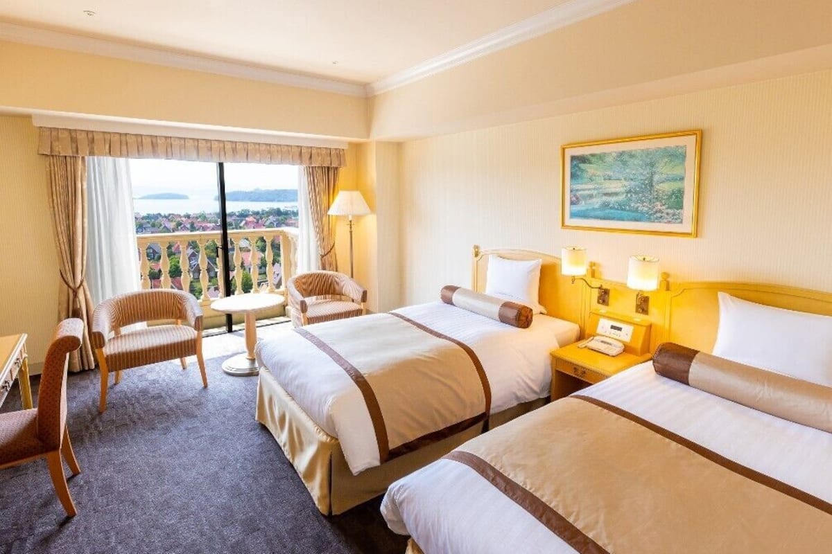 Habitación de hotel con dos camas, un balcón, una mesita y una silla.