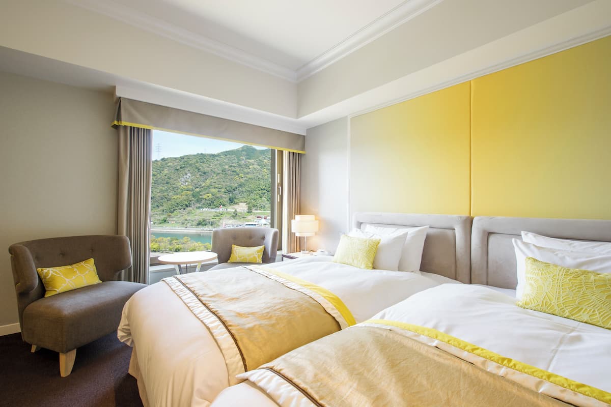 Habitación de hotel con dos camas, una silla, una mesita y vista a un paisaje montañoso.