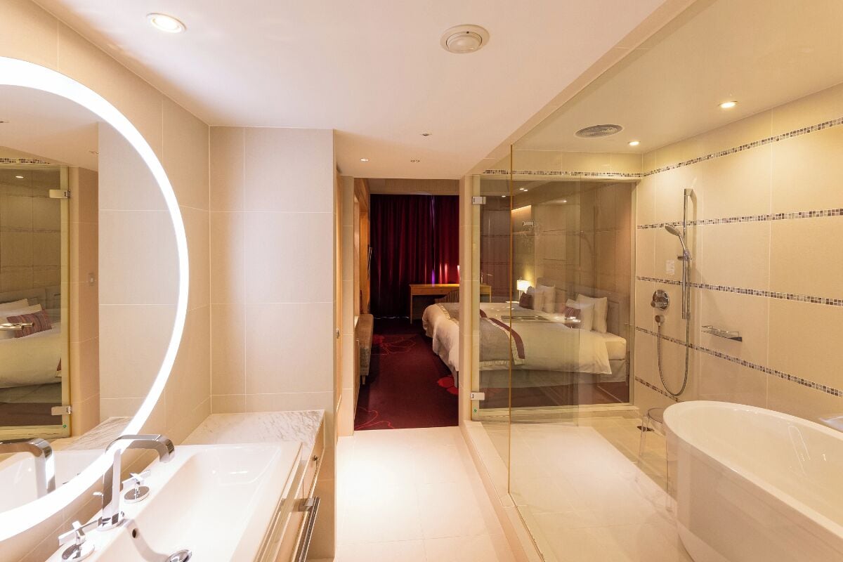 Un baño moderno con un espejo grande, una bañera, zona de ducha y un dormitorio con paredes de cristal.