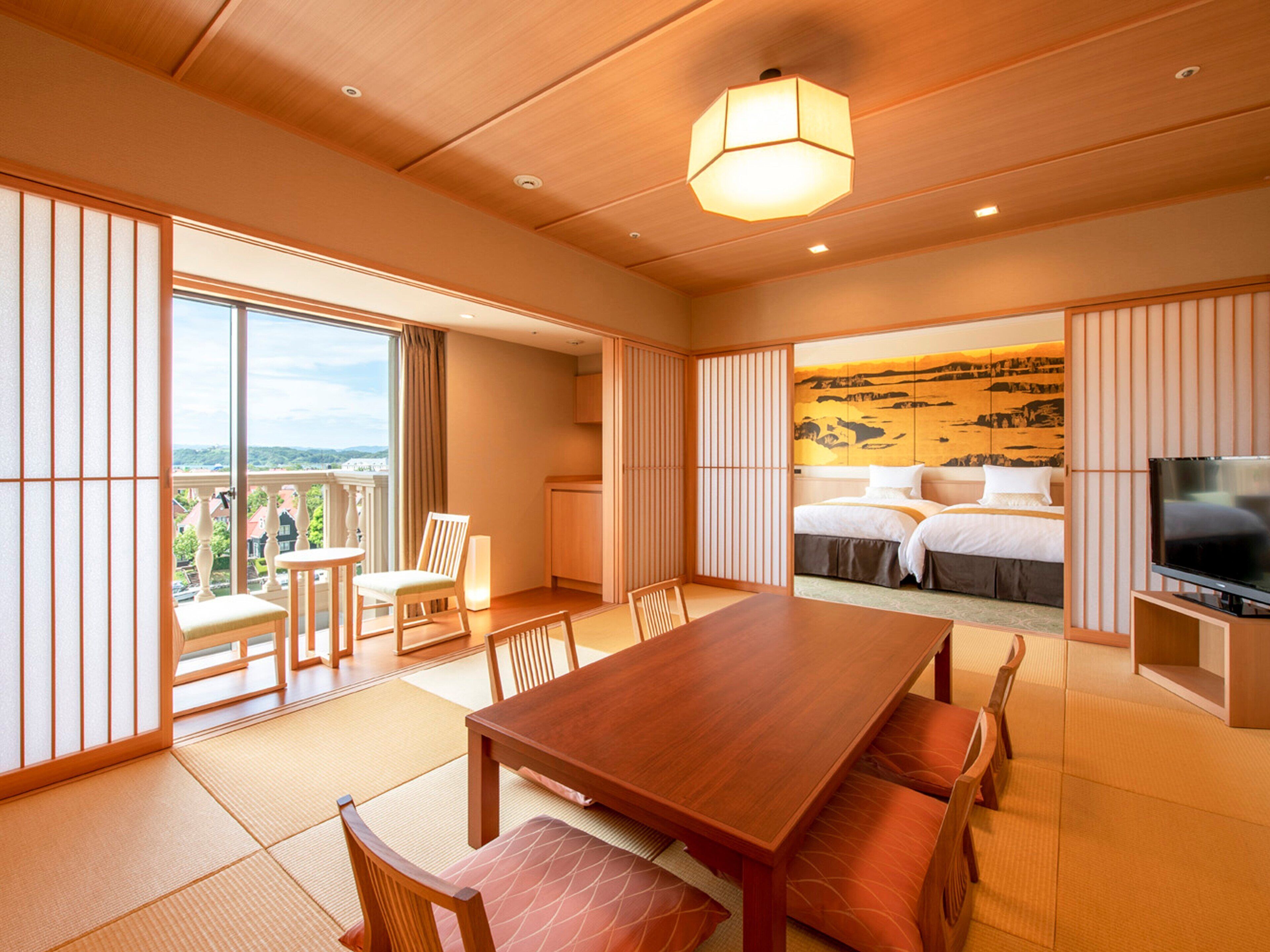 A hotel room with a wooden dining table, chairs, a TV, a bed with a view, and sliding doors leading to a balcony.