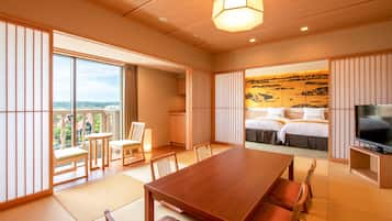 A hotel room with a wooden dining table, chairs, a TV, a bed with a view, and sliding doors leading to a balcony.