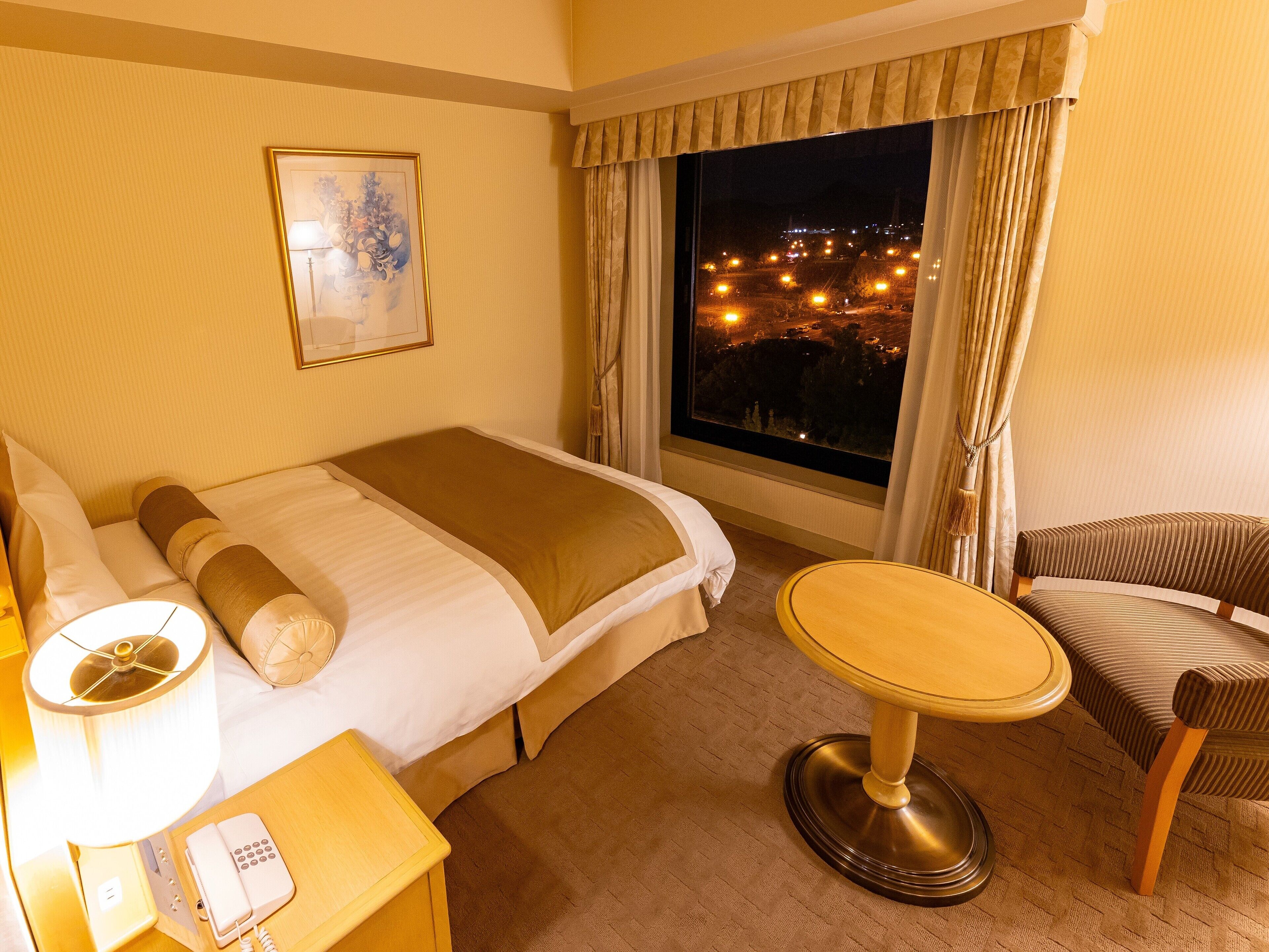 A hotel room with a bed, a bedside table with a lamp and phone, a chair, and a window with a city view at night.