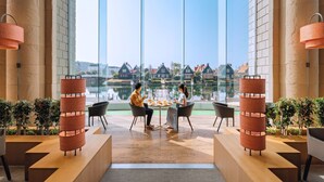 A modern hotel lobby with a large glass window offering a view of traditional houses and a body of water. There are two people seated at a table with a fruit bowl and a bottle. - Hotel Okura JR Huis Ten Bosch (Sasebo)