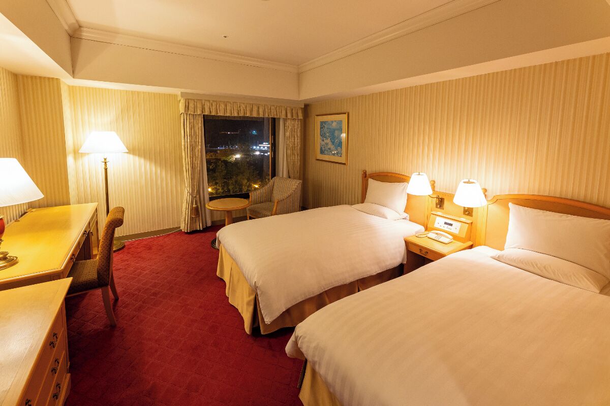 A hotel room with two beds, a desk, a chair, and a small table.