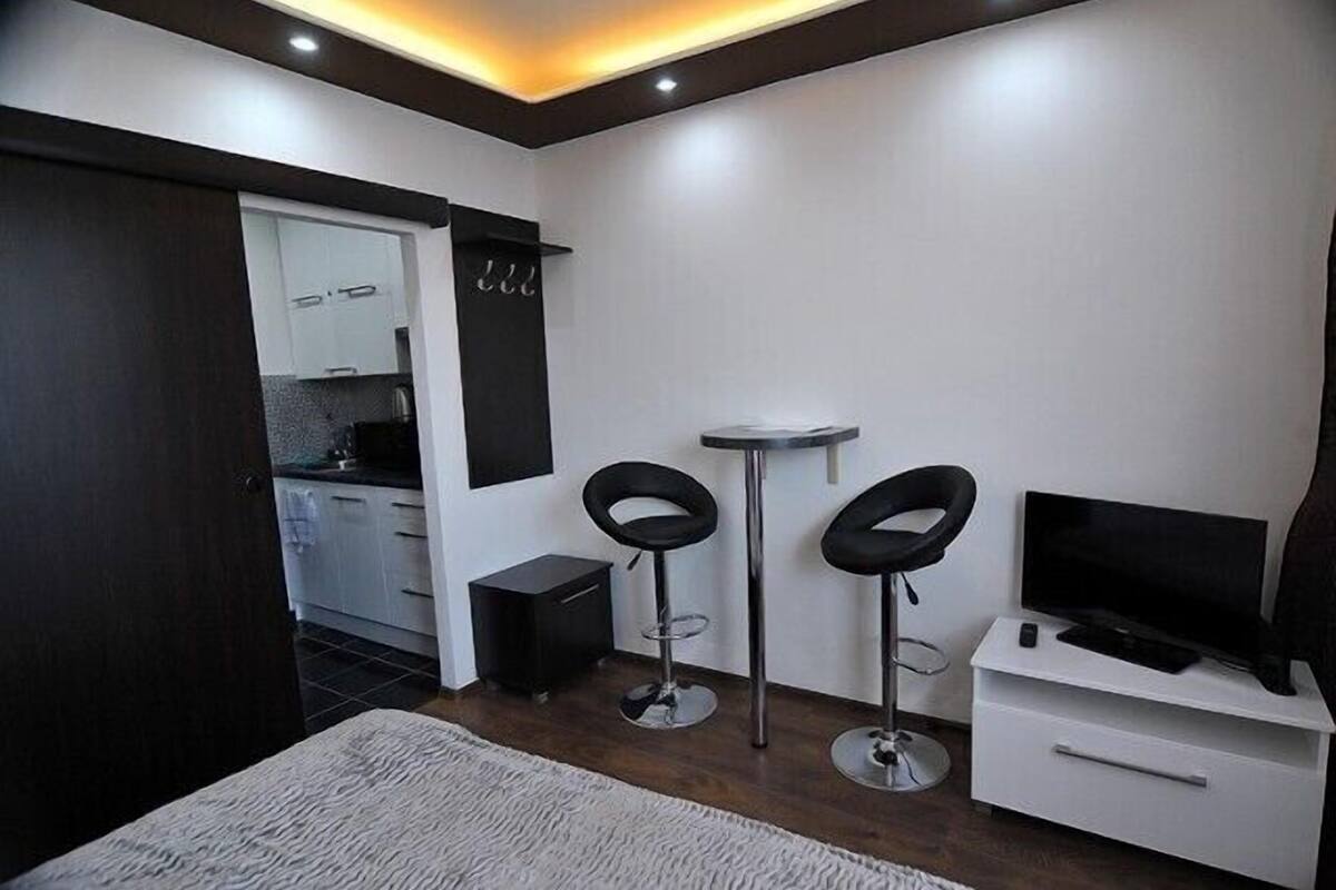 A compact room with a kitchenette, two black bar stools, a TV on a white cabinet, and a door leading to another room.