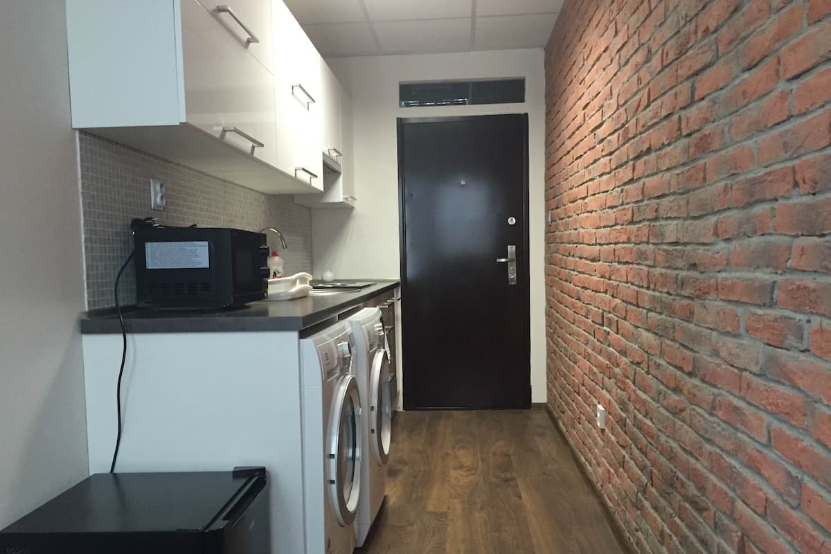 A narrow hallway with a washing machine, a microwave, and a small refrigerator. A door is visible at the end of the hallway.