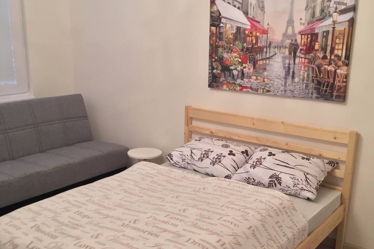 A bedroom with a wooden bed, a grey sofa, a small round table, and a painting of a Paris street scene.