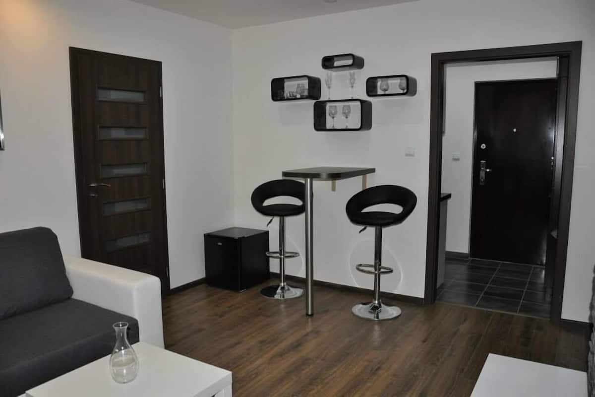 A modern living room with a dark wooden door, a white sofa, a small table with a vase, two black bar stools, a small black speaker, and a wall-mounted wine rack.
