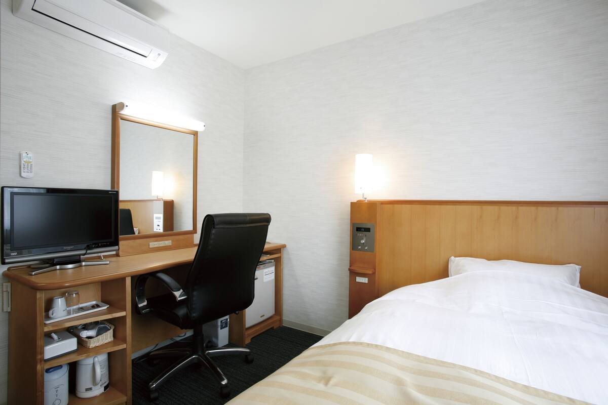 A hotel room with a bed, a desk with a computer, a chair, a small refrigerator, and a mirror.