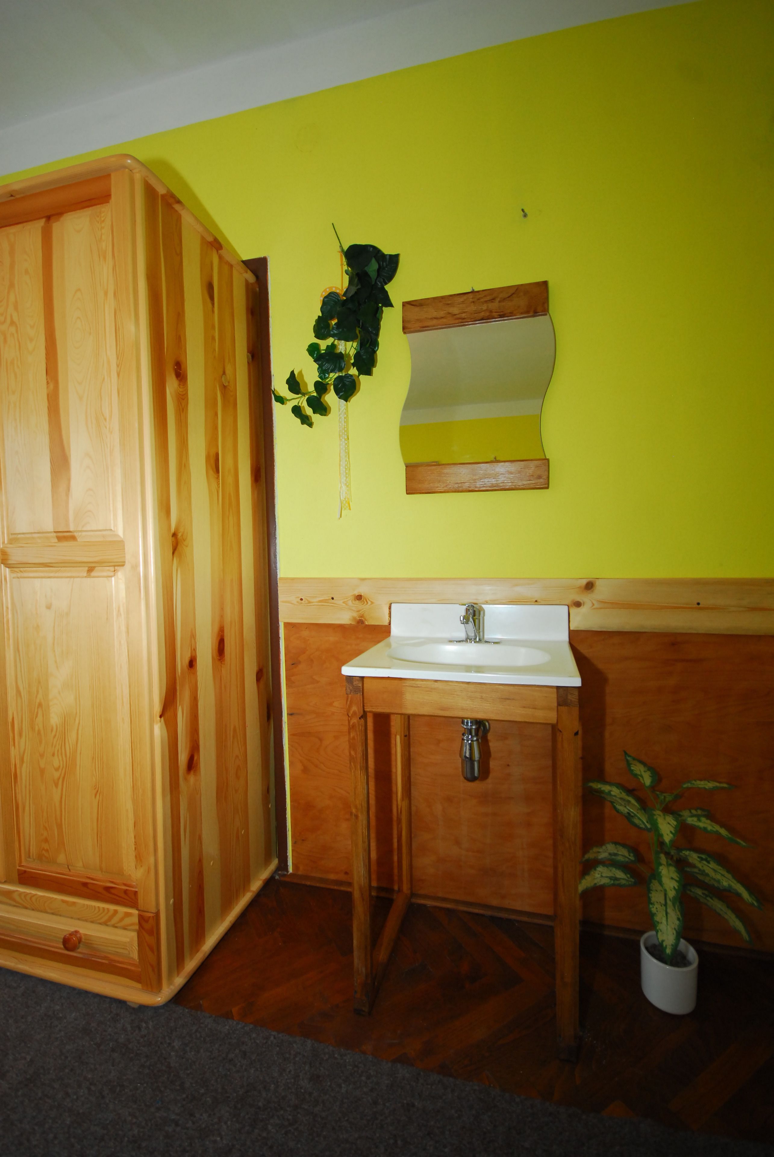 A wooden wardrobe, a white sink with a mirror, and a potted plant in a bathroom with yellow walls.