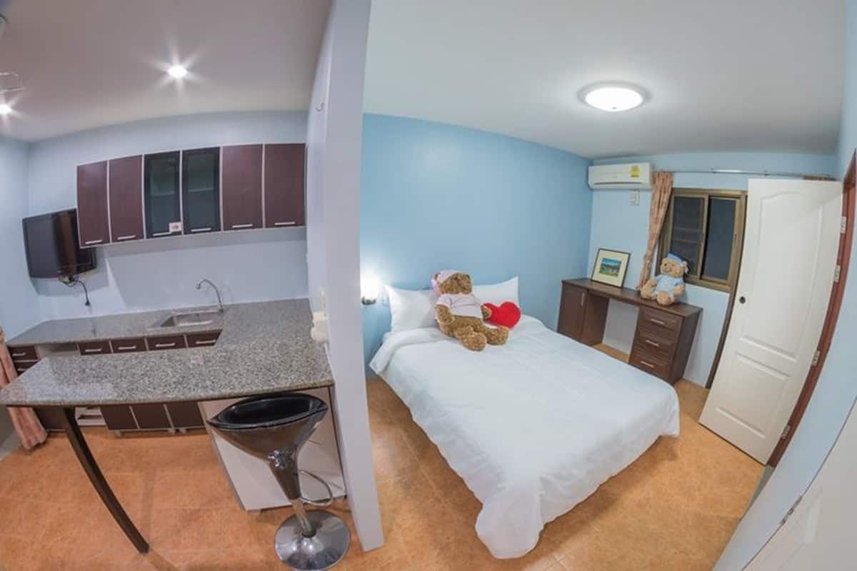 A compact room with a kitchenette, a bed with a teddy bear, and a small dresser.