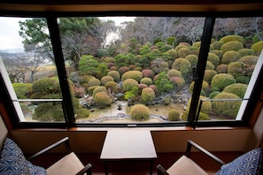 A room with a large window overlooking a meticulously landscaped garden with variously shaped bushes and a small pond. - Shougen Sansou (Tamana)