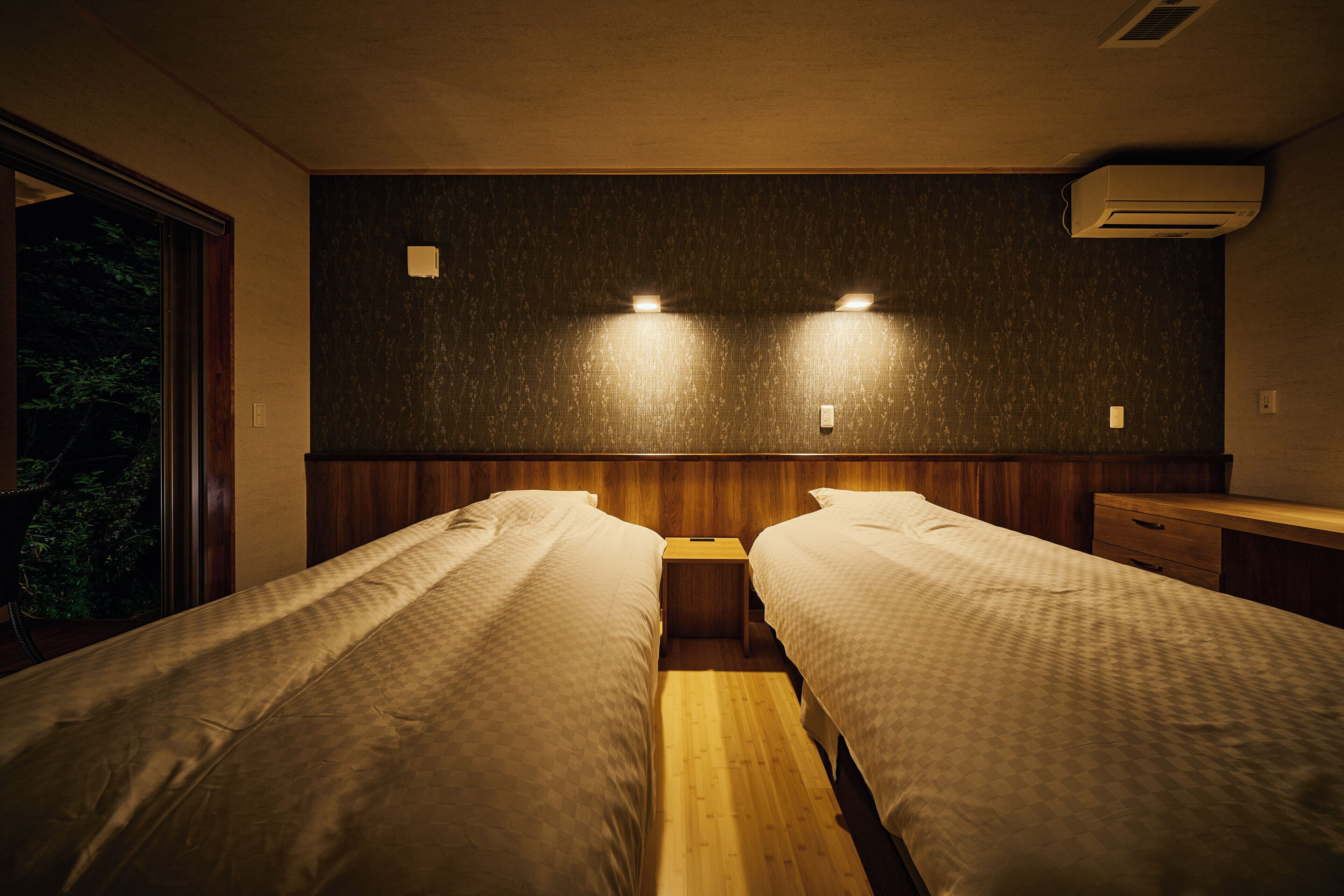 A room with two beds, a wooden floor, and a window with a view of trees.