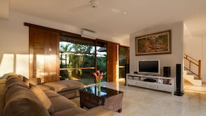 A modern living room with a sectional sofa, a glass coffee table, a flat-screen TV, and a bookshelf. - Balangan Beach Villa (Jimbaran)