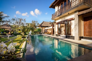 Piscina cercada por plantas tropicais e um edifício de dois andares com varanda.