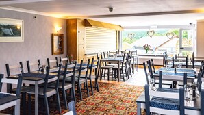 A dining area with multiple tables and chairs, a patterned carpet, and a view of a building with heart-shaped decorations. - The Elan Hotel (Rhayader)