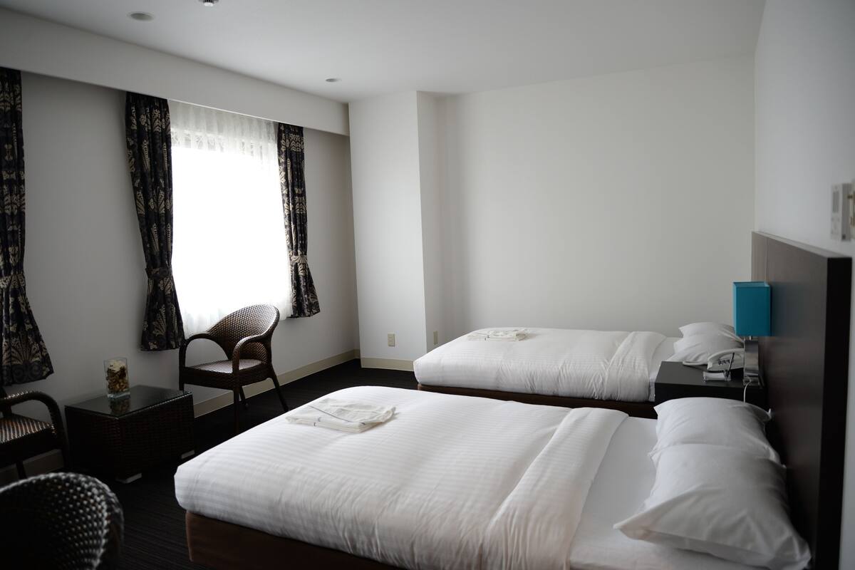 Una habitación de hotel con dos camas, una mesita pequeña, una silla y una mesita de noche con lámpara.