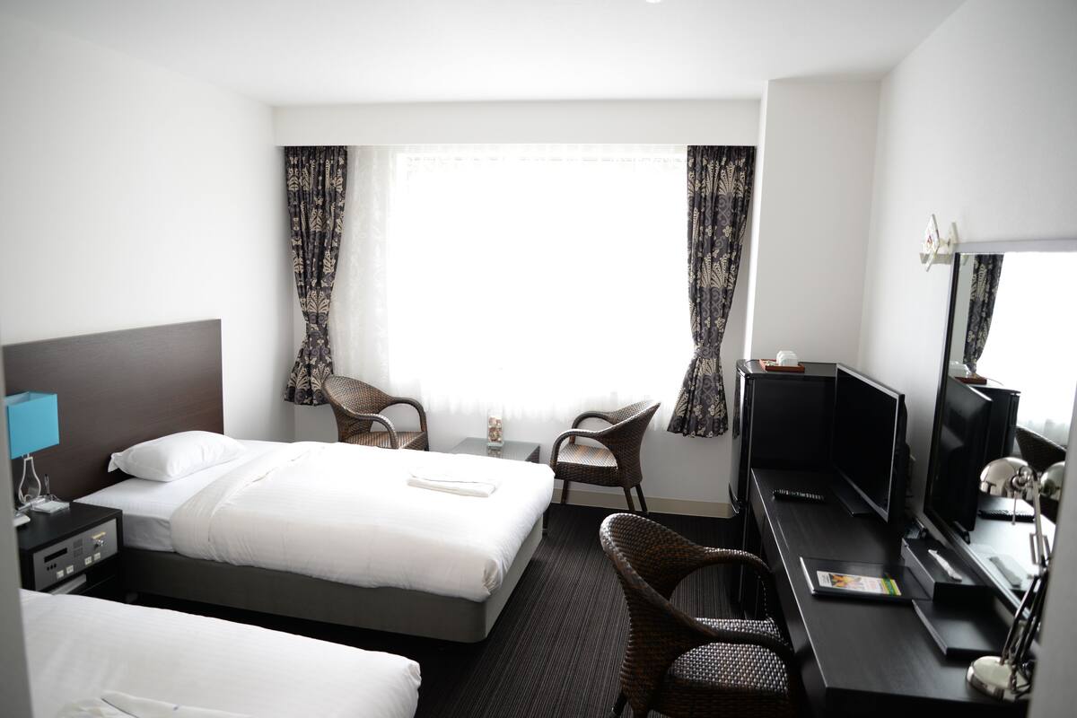 Una habitación de hotel con una cama, dos sillas, un escritorio con computadora, un televisor y una ventana con cortinas.